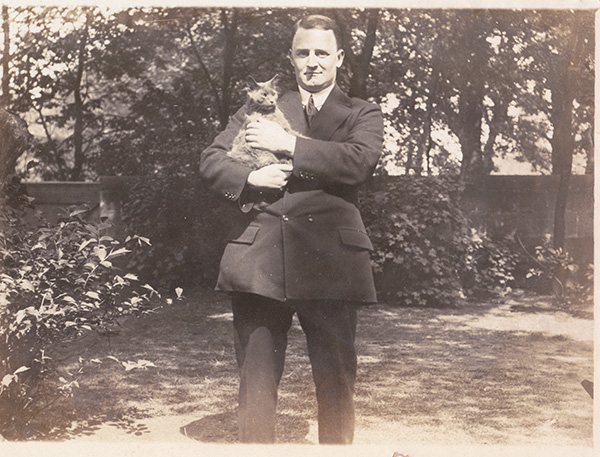 Sepia photo of a man with light skin in a dark suit standing in a garden and holding a cat.